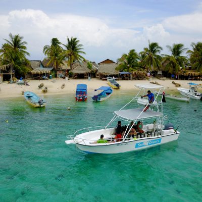 the Sun Divers boat cruises by a small island with fishing boats lined up along the beach and palm trees and huts on the shore. 