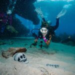 A diver pretends to scream underwater next to a skull and crossbones buried under sand during a halloween event dive in roatan.