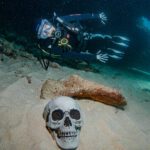 A diver dressed as a skeleton poses next to fake skull and cross bones buried in the sand on a halloween event dive in roatan.