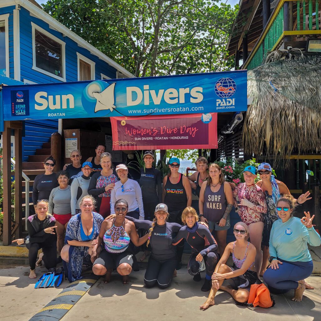 A large group of women is posing for a photo in front of the dive shop Sun Divers in Roatan, Honduras. In the back a flyer reads:"Women's Dive Day".