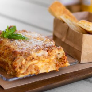 A large portion of cheesy lasagna from Roatan restaurant Drunken Sailor sits on a wooden serving platter with a small brown bag of bread on the side.