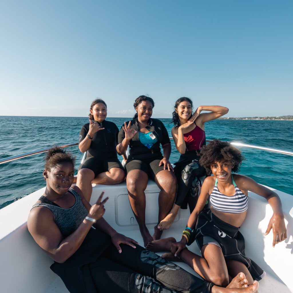 Young islander girls from Roatan sit on the bow of the boat, smiling and giving the okay sign after a dive.