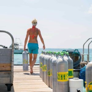 A woman who has grabbed a nitrox tank from two rows on a dock is carrying it to the end of the dock.