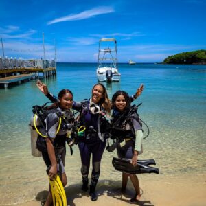Two newly certified girls who received the ROA GIRLS WHO DIVE scholarship are standing at the beach in their SCUBA gear with their instructor.