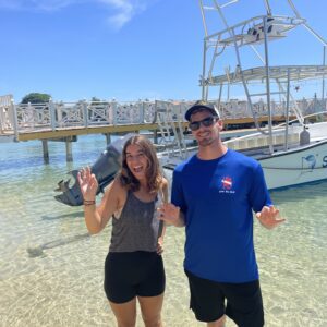 A guy and girl in beach clothes stand on the shore in front of a boat flashing the peace sign and chaka sign.They are part of the Sun Divers work exchange program in Roatan.