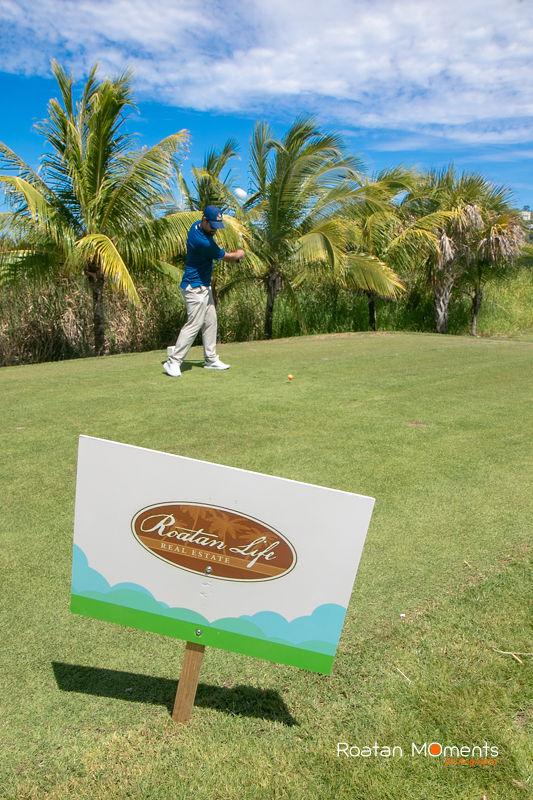 Golfer swings on tropical golf course with Roatan Life sponsor sign in the foreground