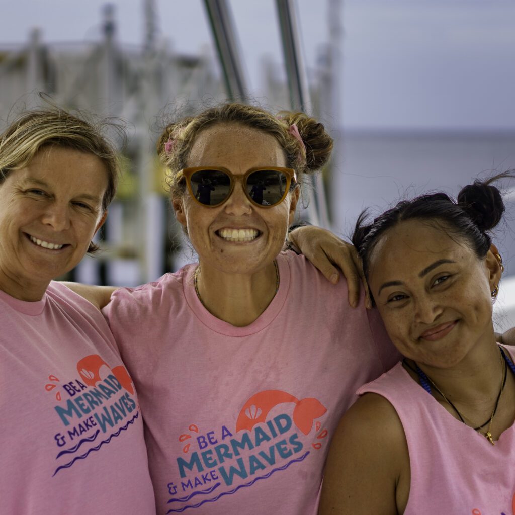 Three women gather for a photo on the boat before going out diving at a PADI women's dive day event in Roatan. They are wearing pink shirts that say "Be a Mermaid and Make waves."