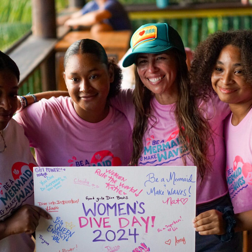 Sun Divers owner Natalie Shuman photographed with 3 scuba scholarship recipients. They are holding a sign that says Women's Dive Day 2024.