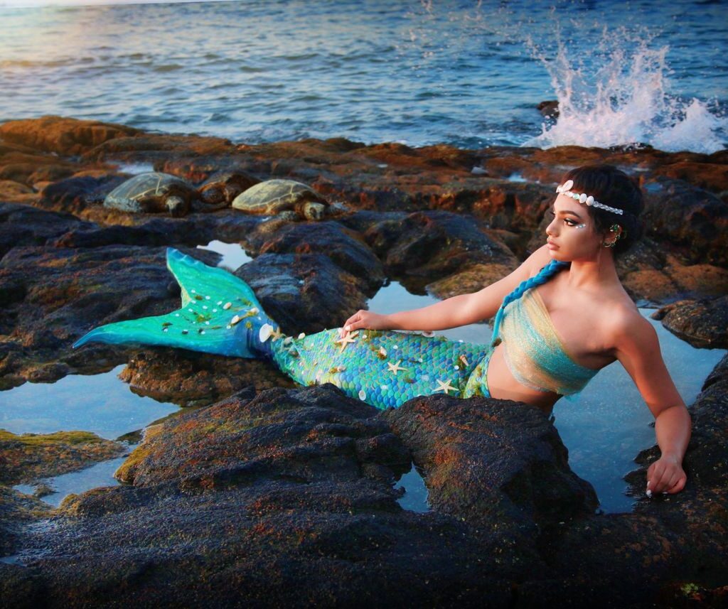 A women dressed as a mermaid poses on coastal rocks while the ocean crashes behind her creating ocean spray