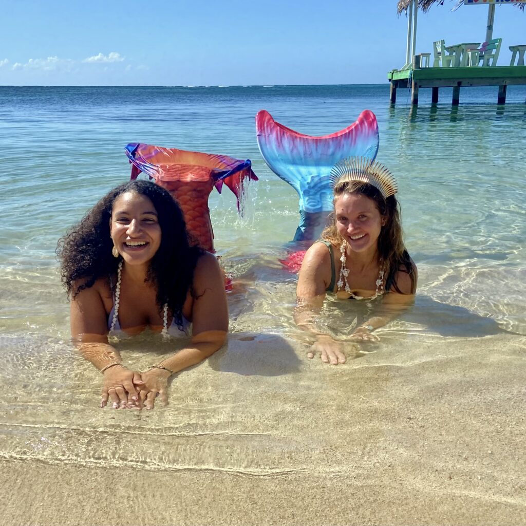 Two women lay on the shore with their mermaid tails flipped in the air. 