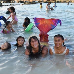 A PADI Mermaid instructor surrounded by young girls flips her tail while posing on the beach.