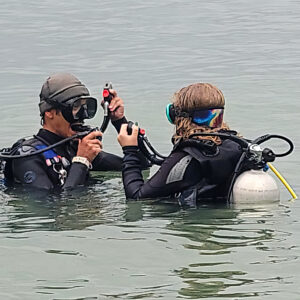 An scuba instructor shows a student how to insert the regulator and deflate the bcd to go underwater in confined water. 