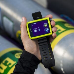 Photo of a hand holding a dive computer with scuba tanks in the background.